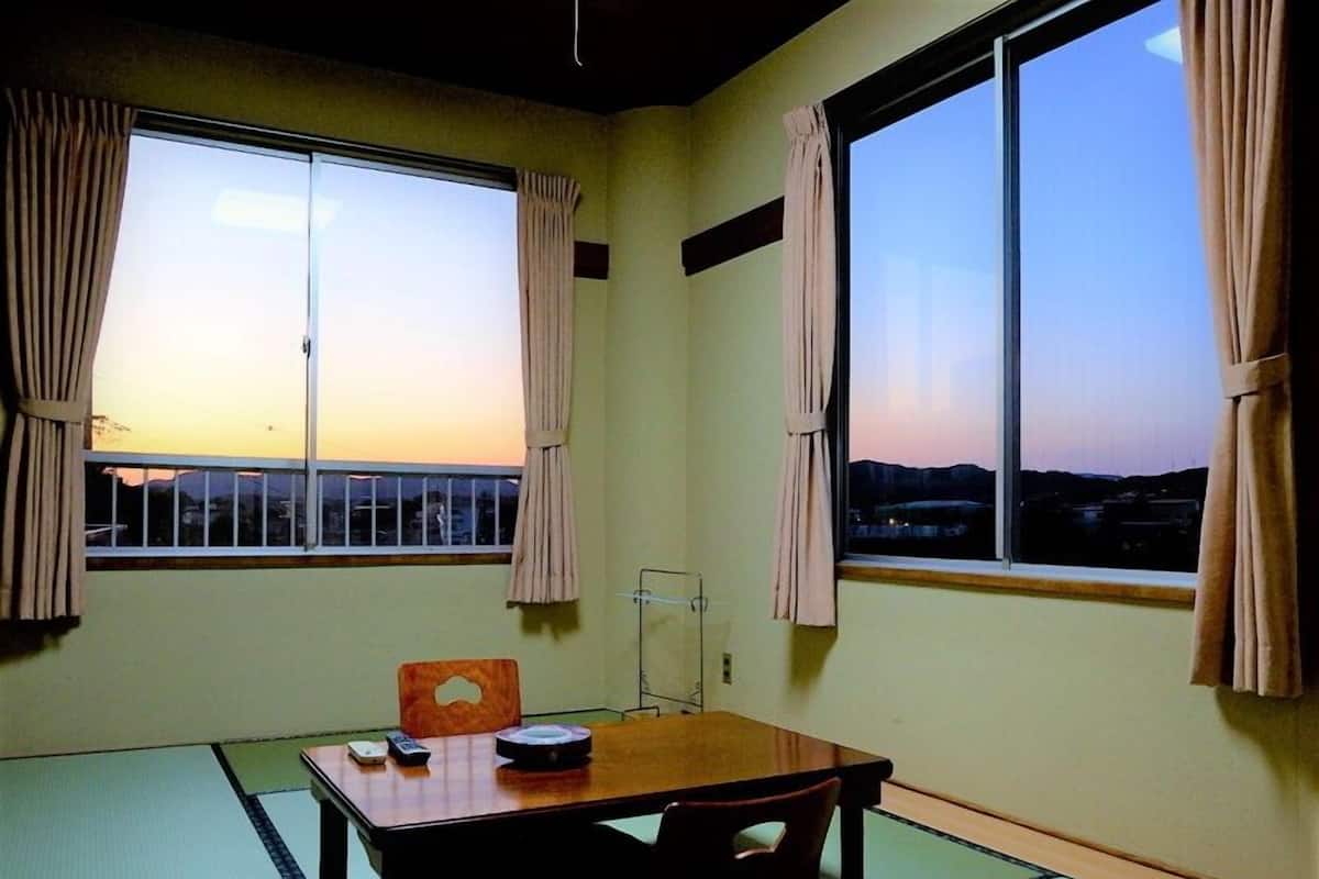 A room with tatami mat flooring, a wooden table with a remote control, and a view of a sunset through large windows.