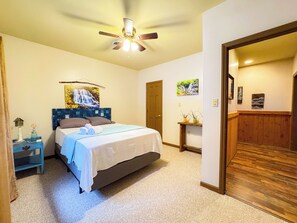 Une chambre à coucher comprenant un lit, une table de chevet, un ventilateur de plafond et une porte donnant sur une autre pièce.