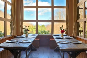 Ein Essbereich mit Holztischen, gedeckt für vier Personen, großen Fenstern und Blick auf Bäume und Himmel.