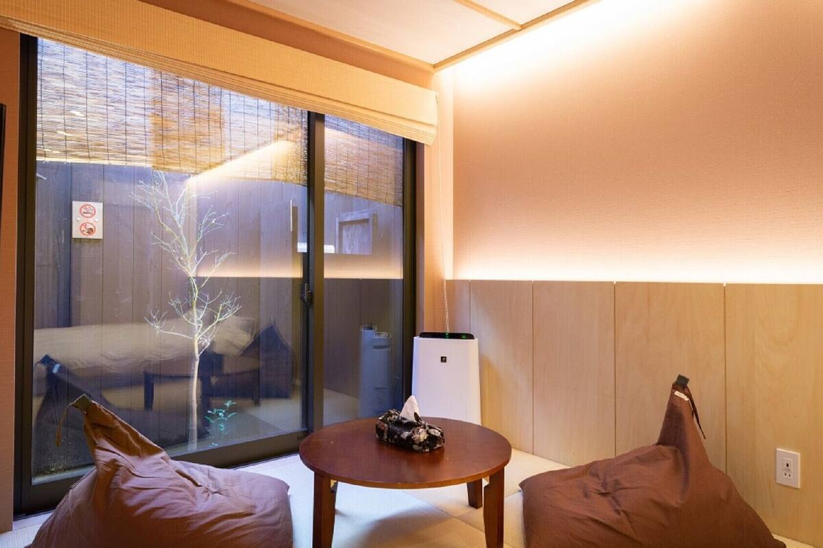 Quarto com porta de vidro deslizante para uma varanda, uma pequena mesa com um objeto decorativo e duas áreas de estar com almofadas.