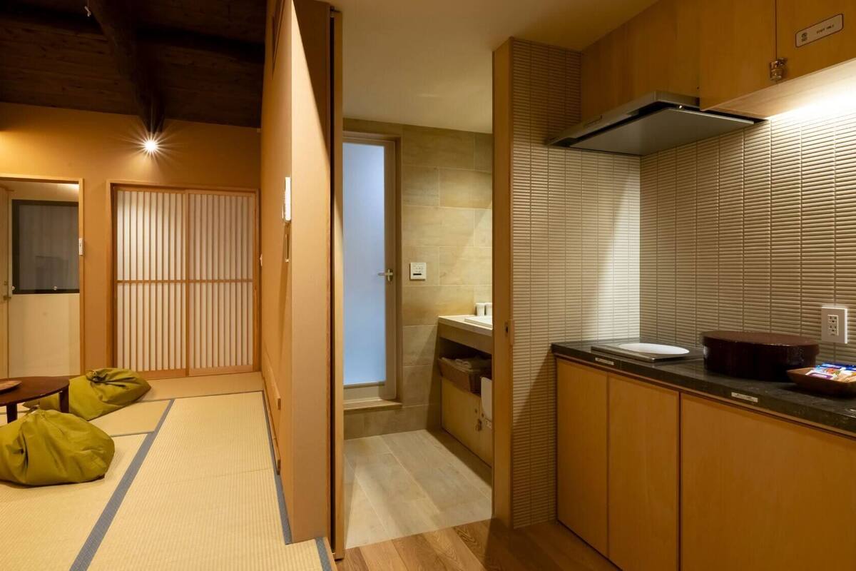 Um quarto japonês tradicional com piso de tatame, porta de shoji, uma pequena mesa com almofada e uma área de cozinha com pia e bancada.