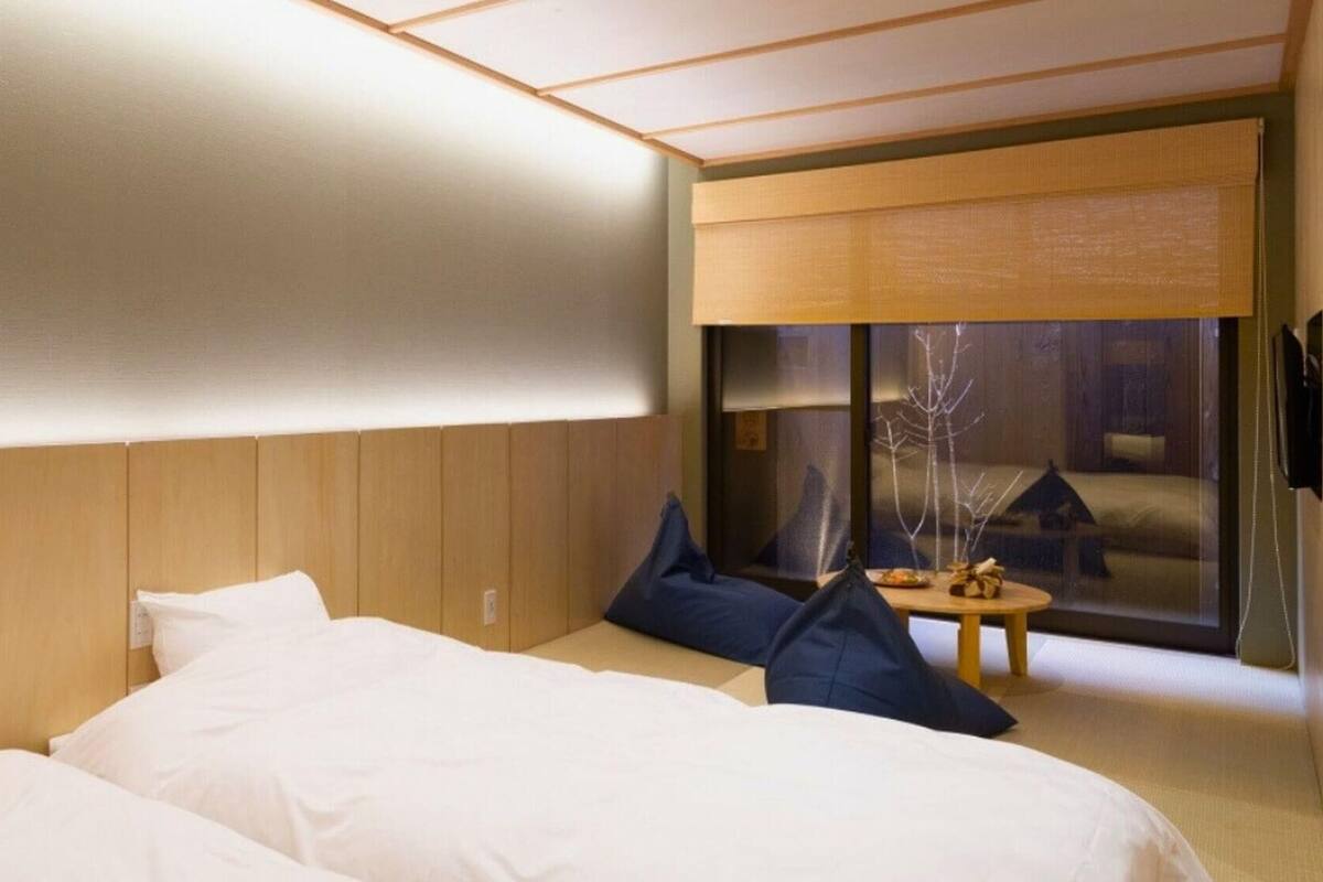 Quarto de hotel com uma cama grande, uma cama menor, uma mesa de trabalho e vista para um edifício através de uma janela.