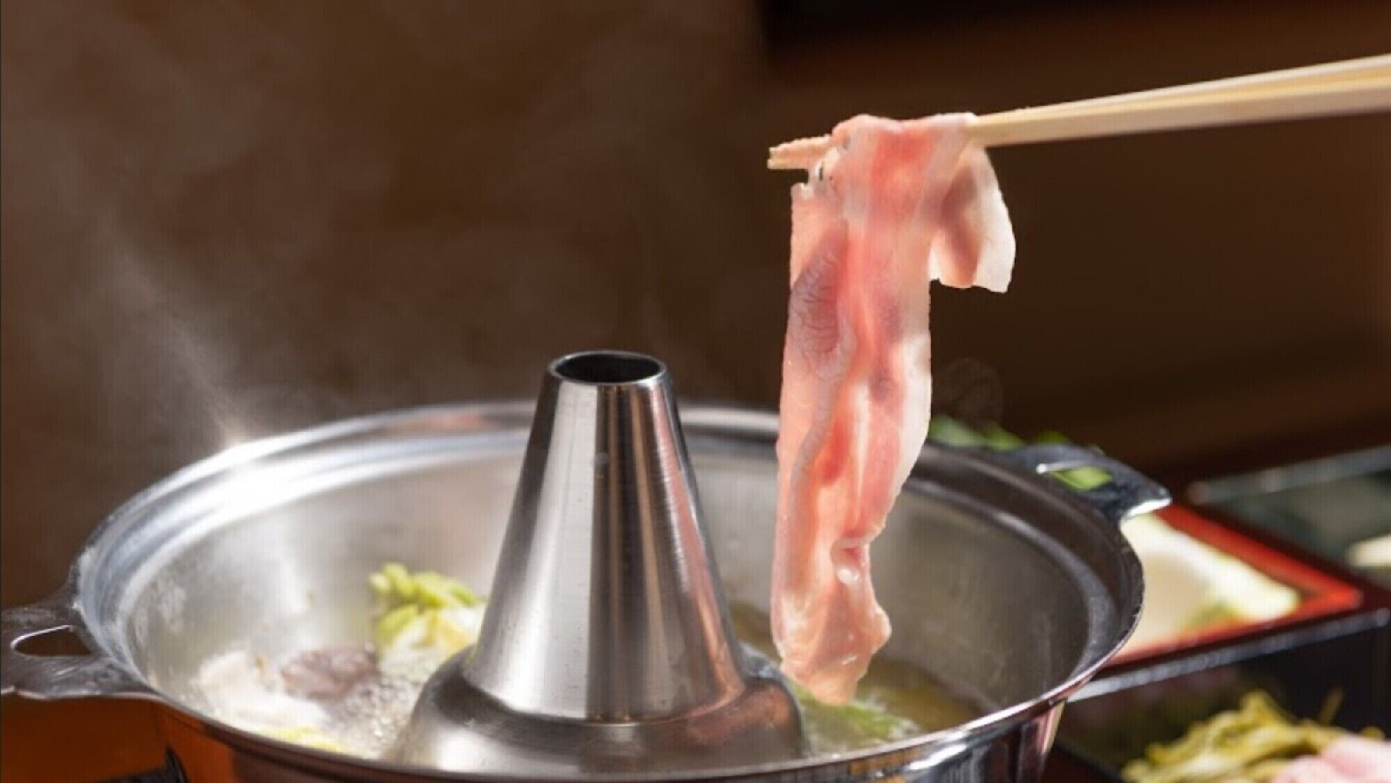 A hot pot with a metal steamer insert and a piece of meat being held by chopsticks.