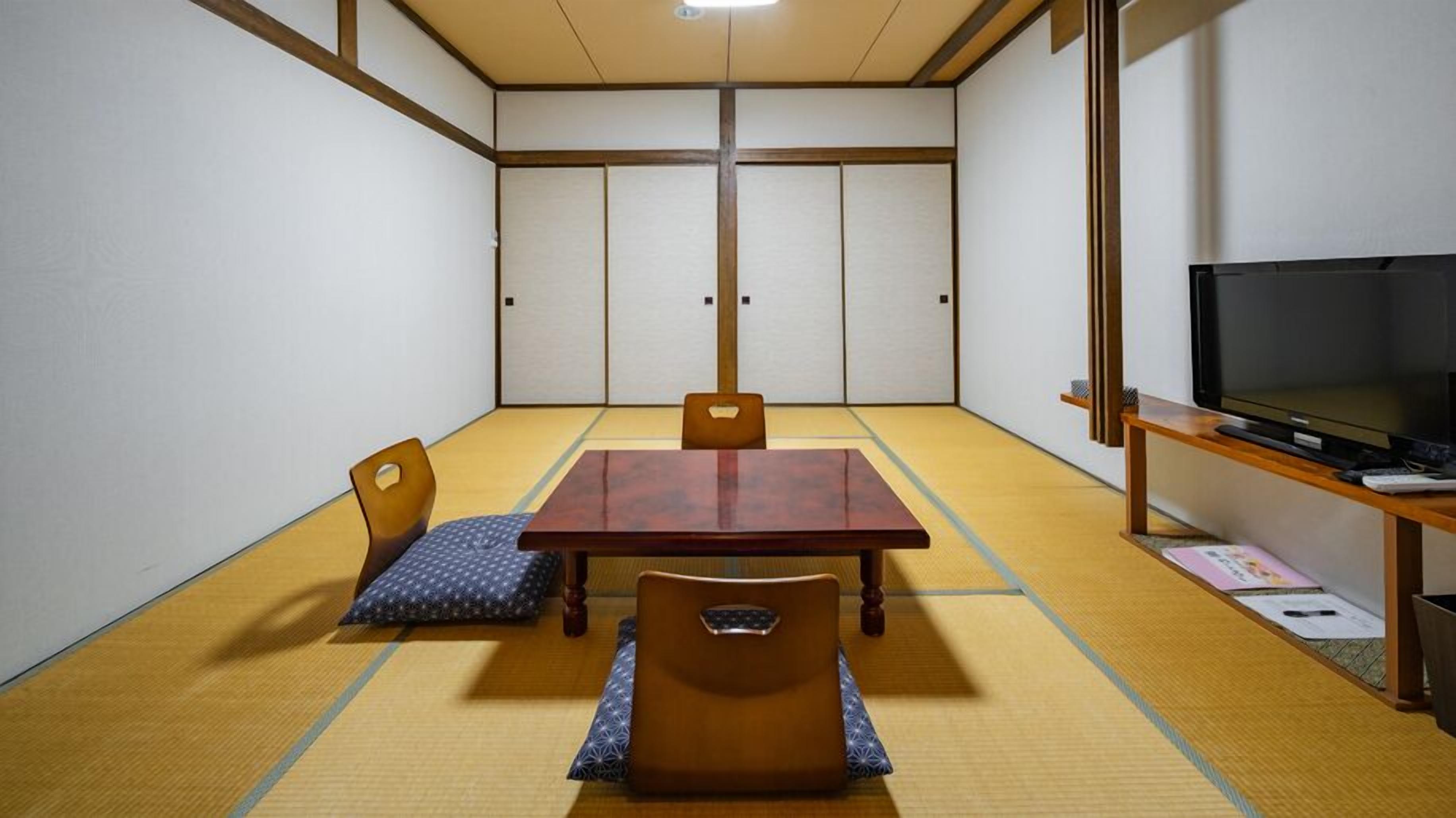 Una habitación con tatamis, una mesa de madera, sillas, un televisor y un estante pequeño.