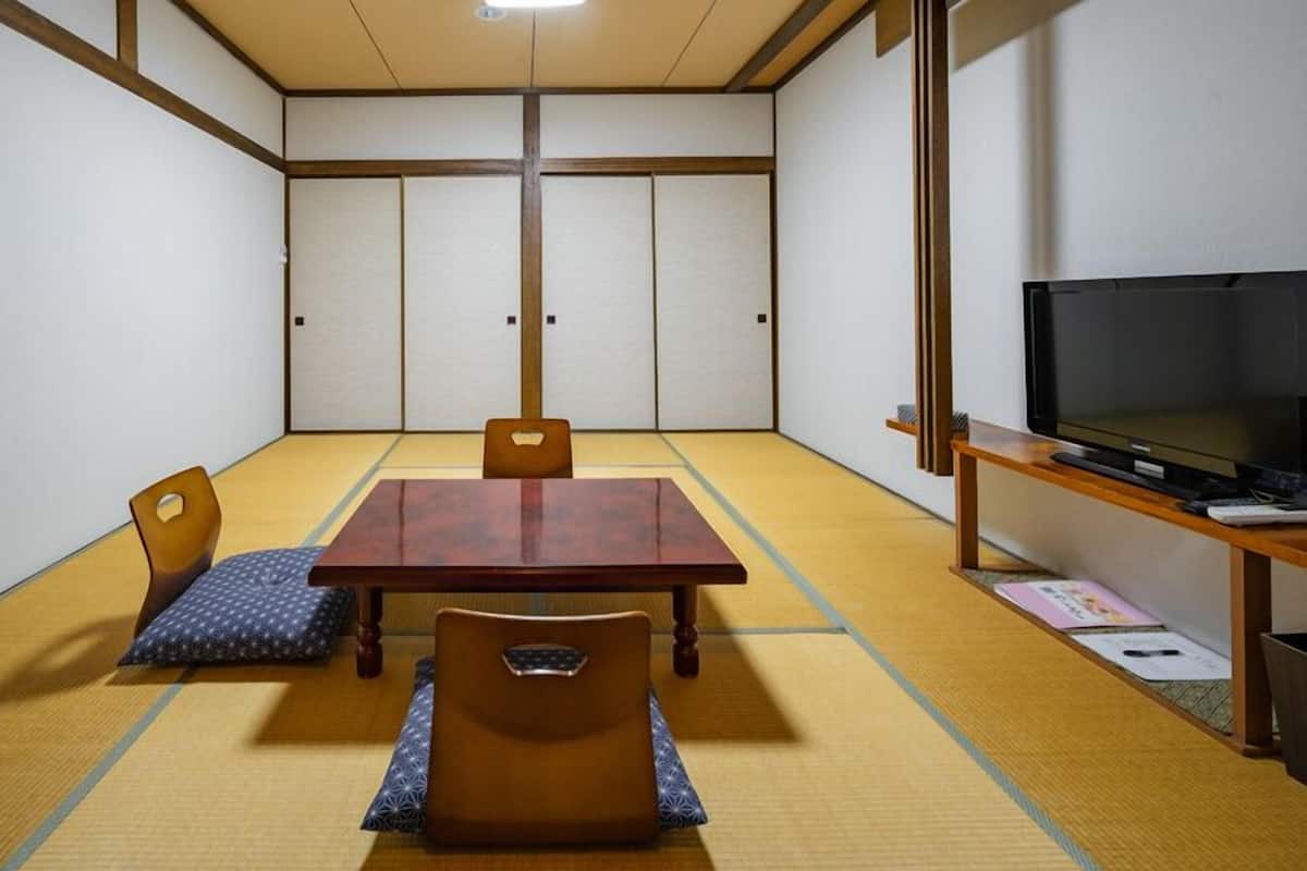 Une pièce avec un sol recouvert de tatamis, une table en bois, des chaises, un téléviseur et une petite étagère.