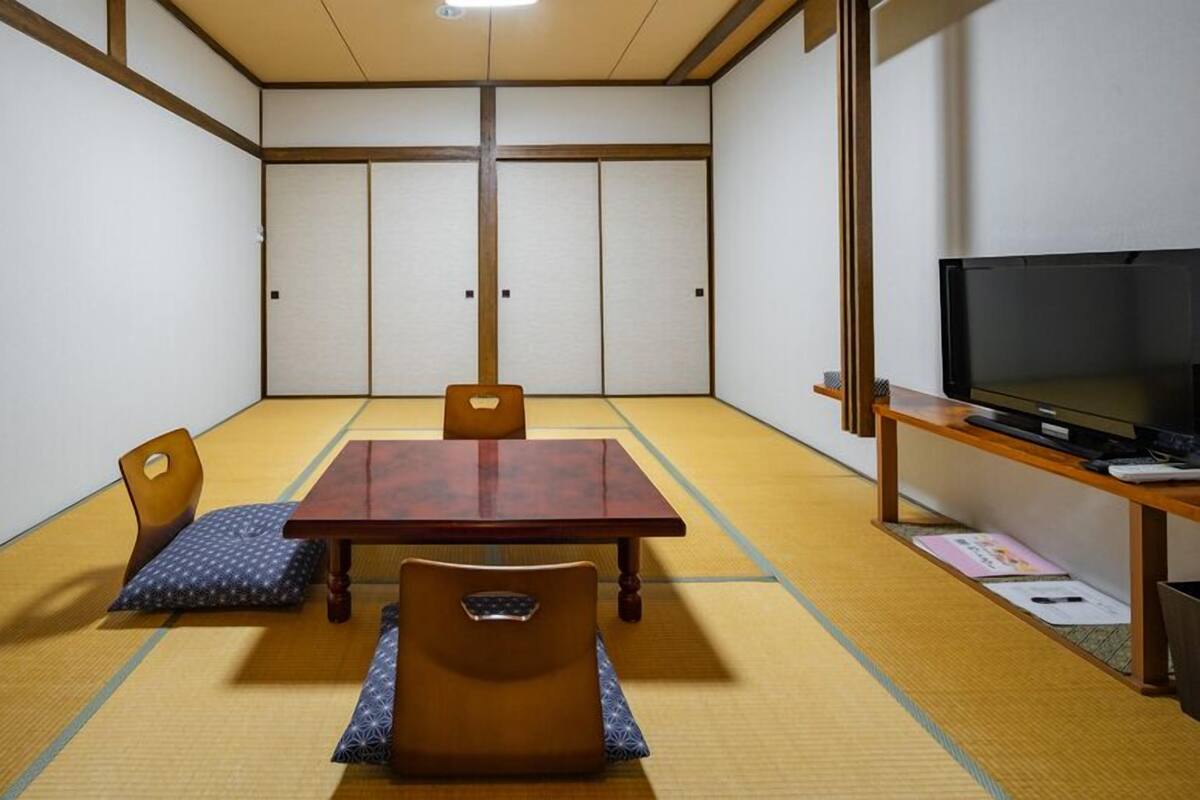Una habitación con tatamis, una mesa de madera, sillas, un televisor y un estante pequeño.
