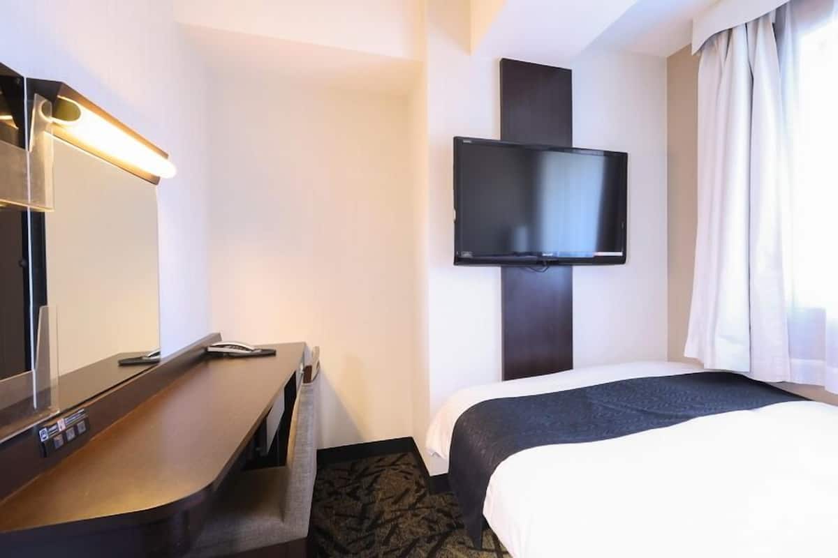 A hotel room with a bed, a desk, a flat-screen TV mounted on the wall, and a window with curtains.