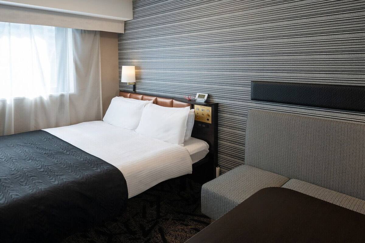 A hotel room with a bed, bedside table, lamp, and a view of the outdoors.