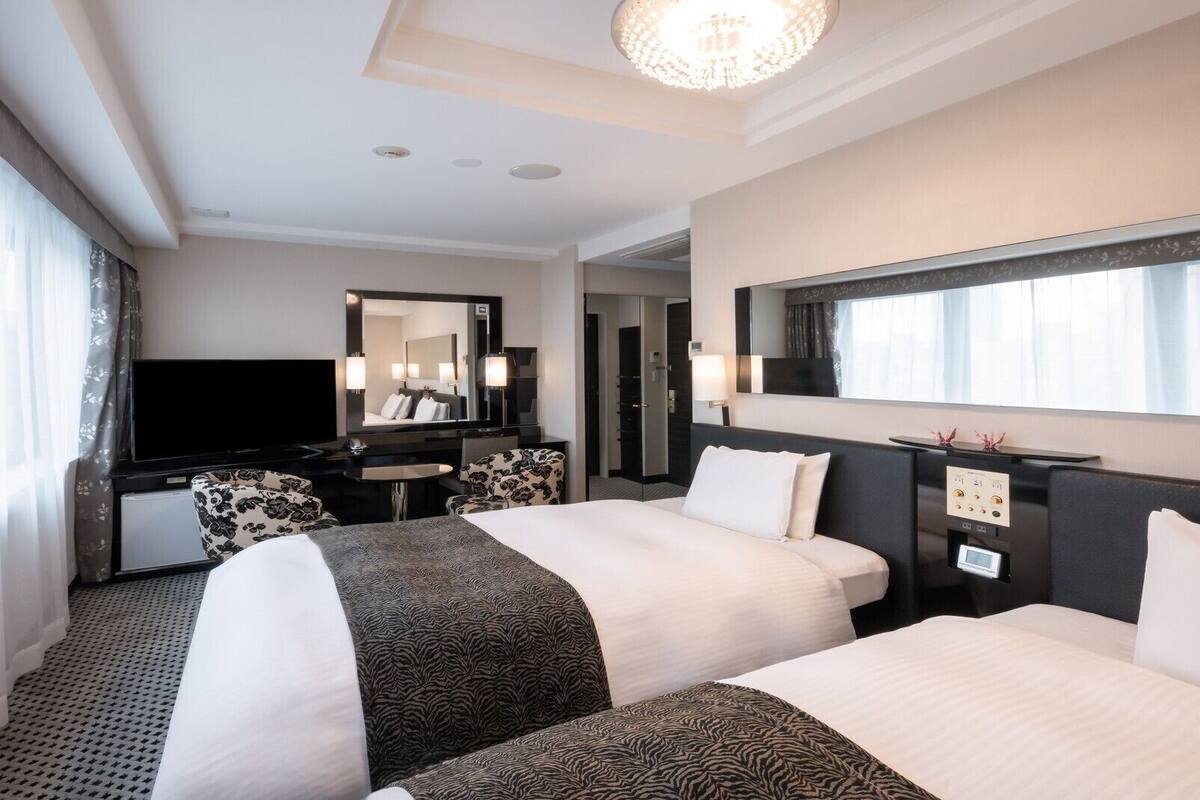 A hotel room with two beds, a TV, a desk, a mirror, and a chandelier.