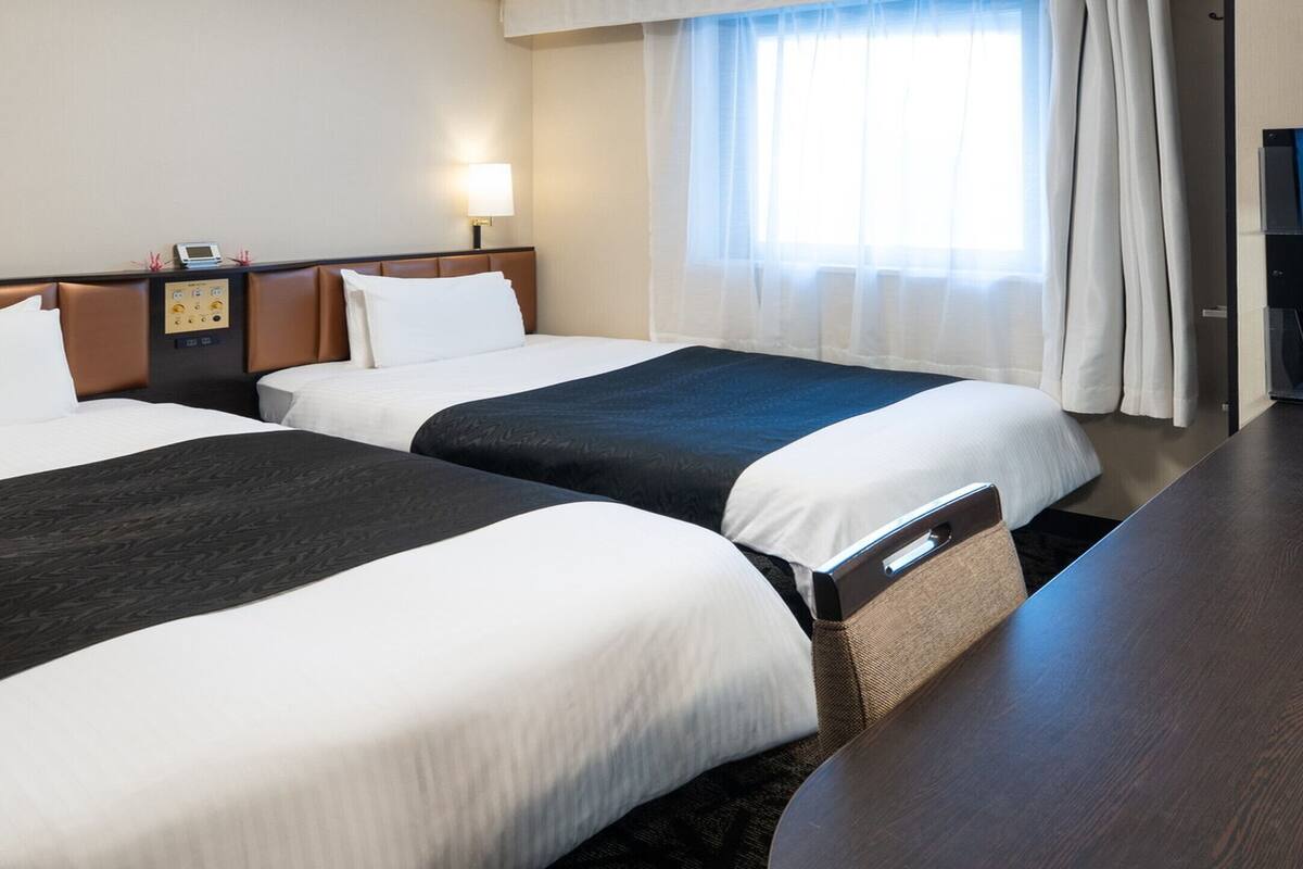 A hotel room with two beds, a desk, a lamp, a clock, and a window with curtains.