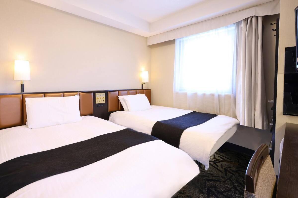 A hotel room with two beds, a desk, a chair, a television, and a window with curtains.