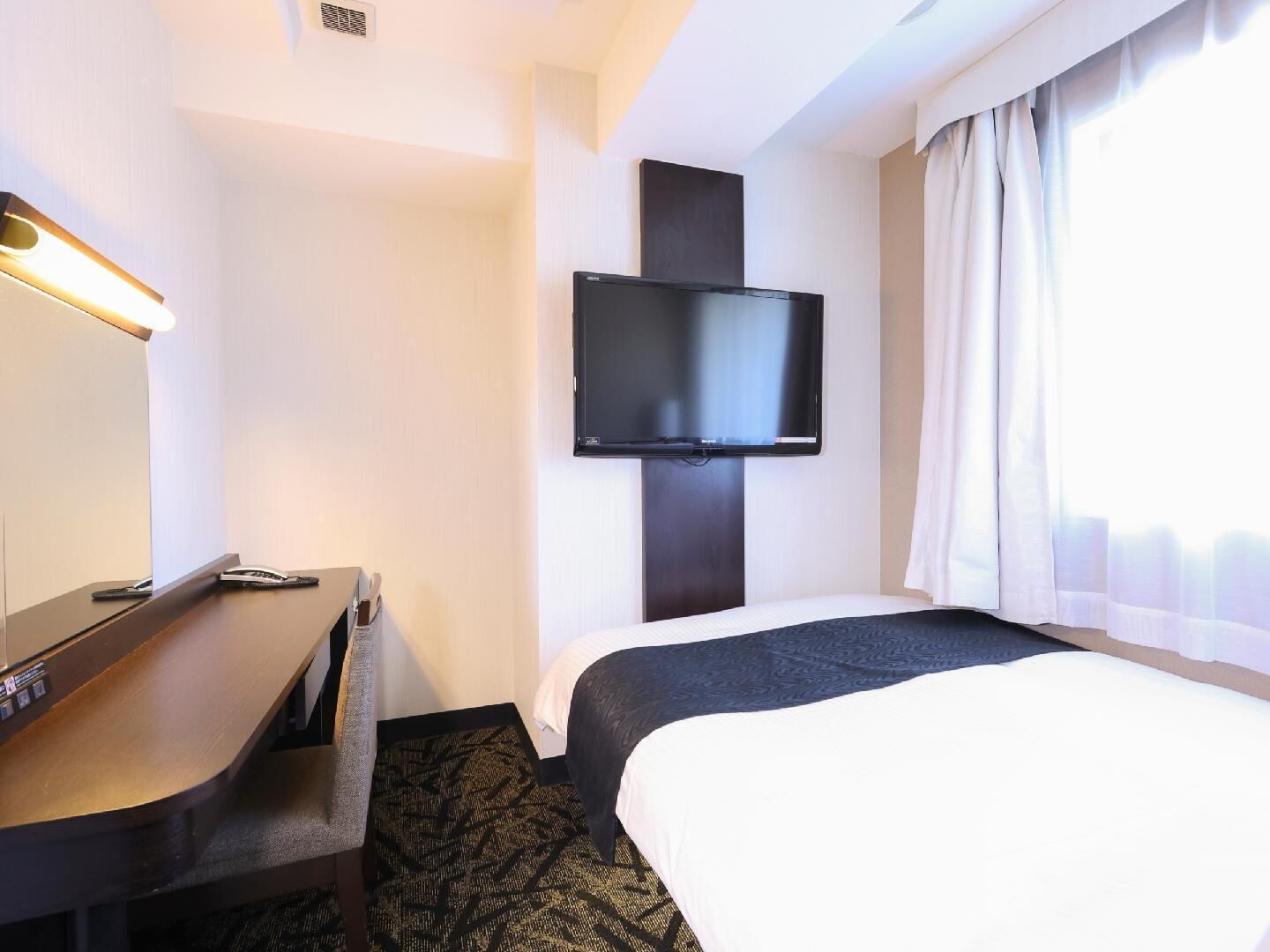 Una camera d'albergo con un letto, una scrivania, una TV a schermo piatto montata a parete e una finestra con tende.