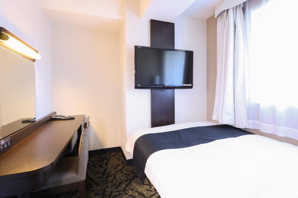 A hotel room with a bed, a desk, a flat-screen TV mounted on the wall, and a window with curtains.