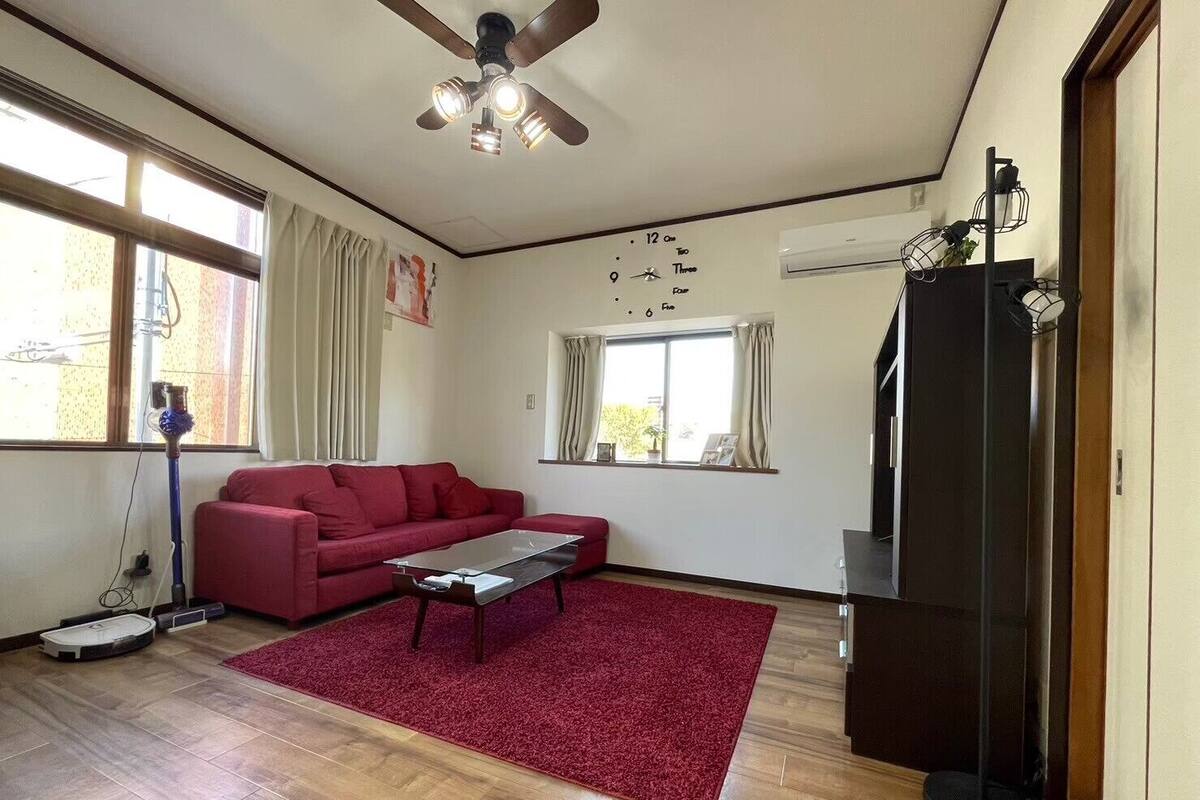 A living room with a red sofa, a coffee table, a red rug, a ceiling fan, and a large window with curtains.