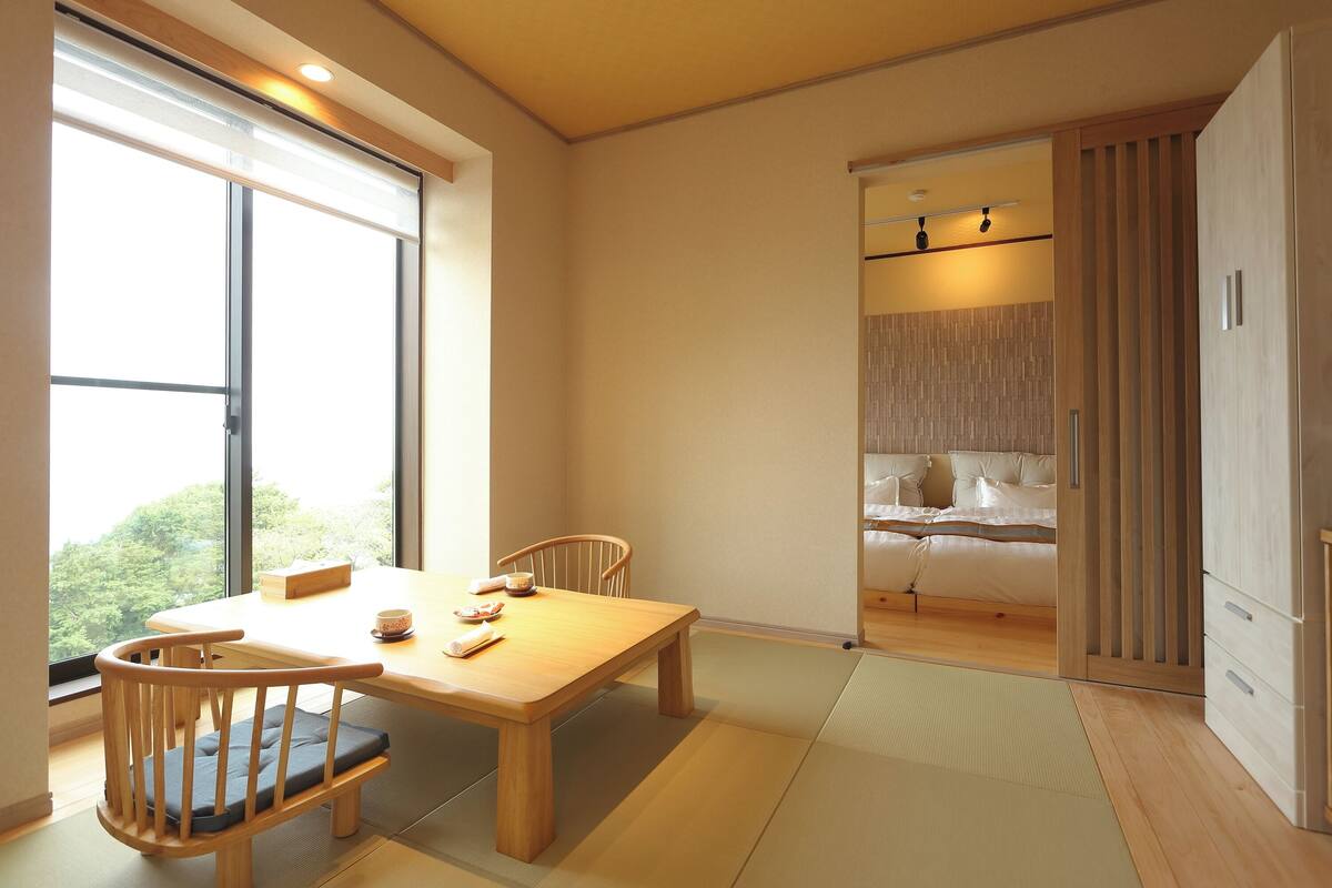 A room with a wooden dining table, chairs, a bed with a view, and a sliding door leading to another room.