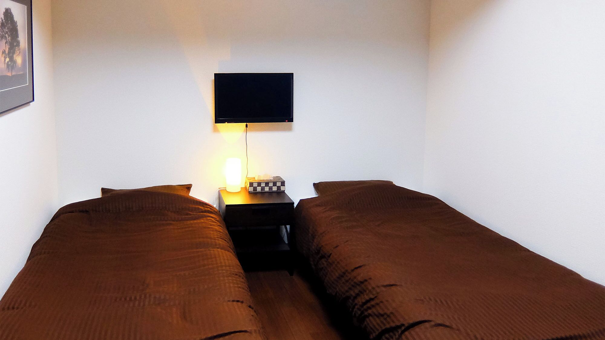 A room with two single beds, a bedside table with a lamp, and a flat-screen TV mounted on the wall.