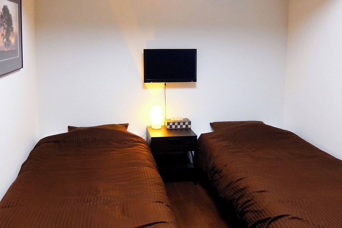 A room with two single beds, a bedside table with a lamp, and a flat-screen TV mounted on the wall.