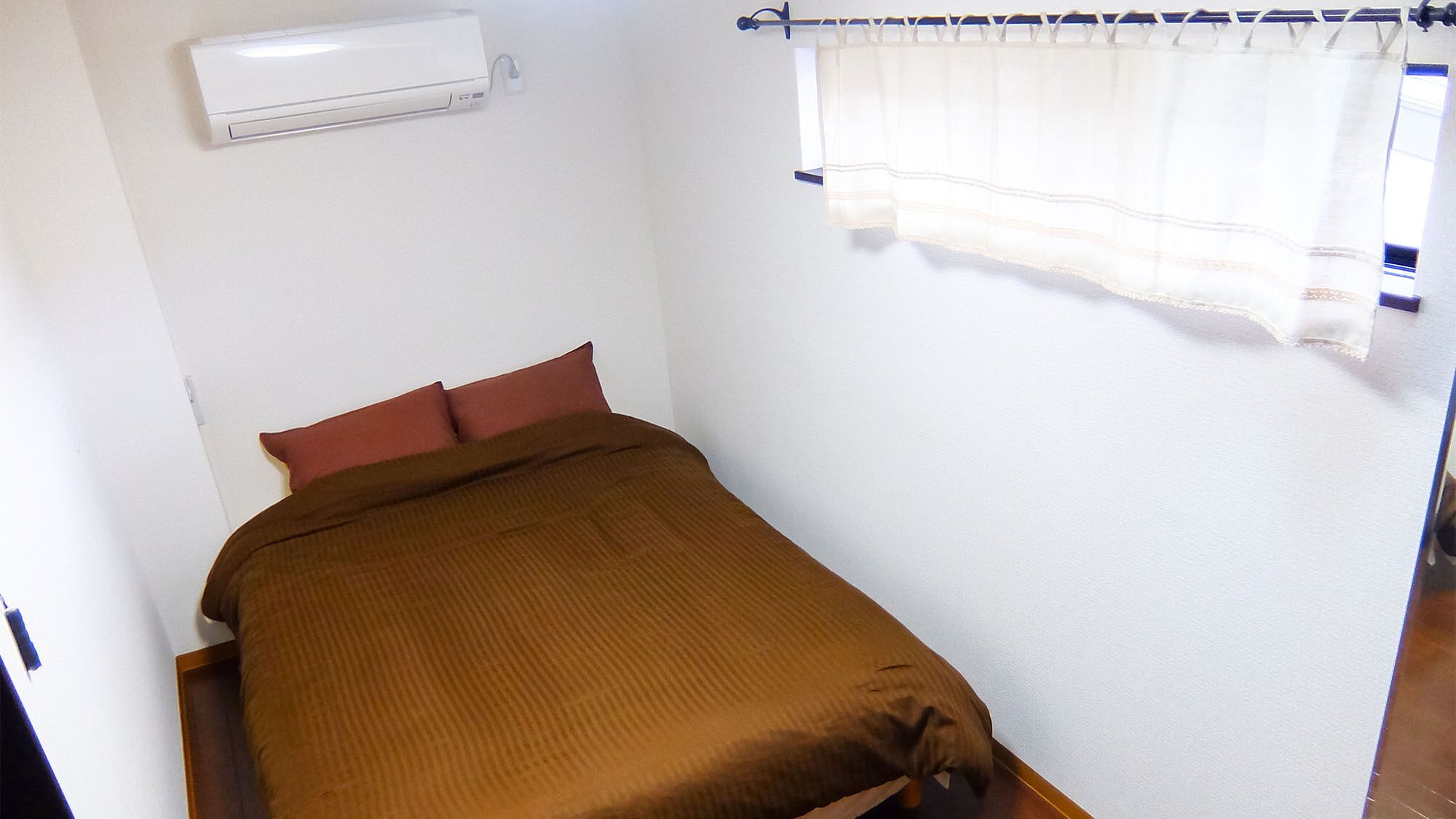 A single bed with a brown blanket, a white wall, and a window with a curtain.