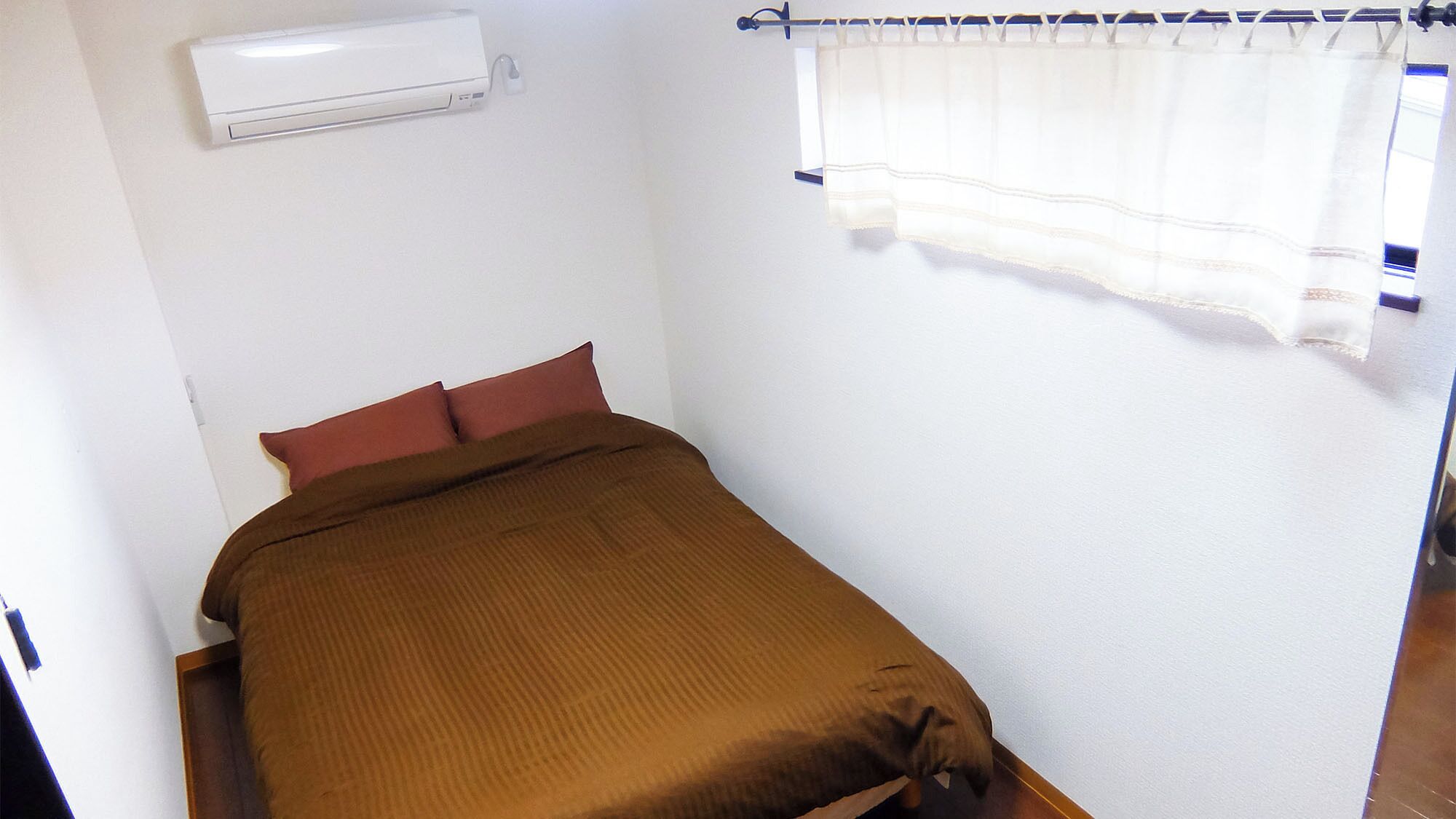 A single bed with a brown cover, a white air conditioner mounted on the wall, and a window with a white curtain.