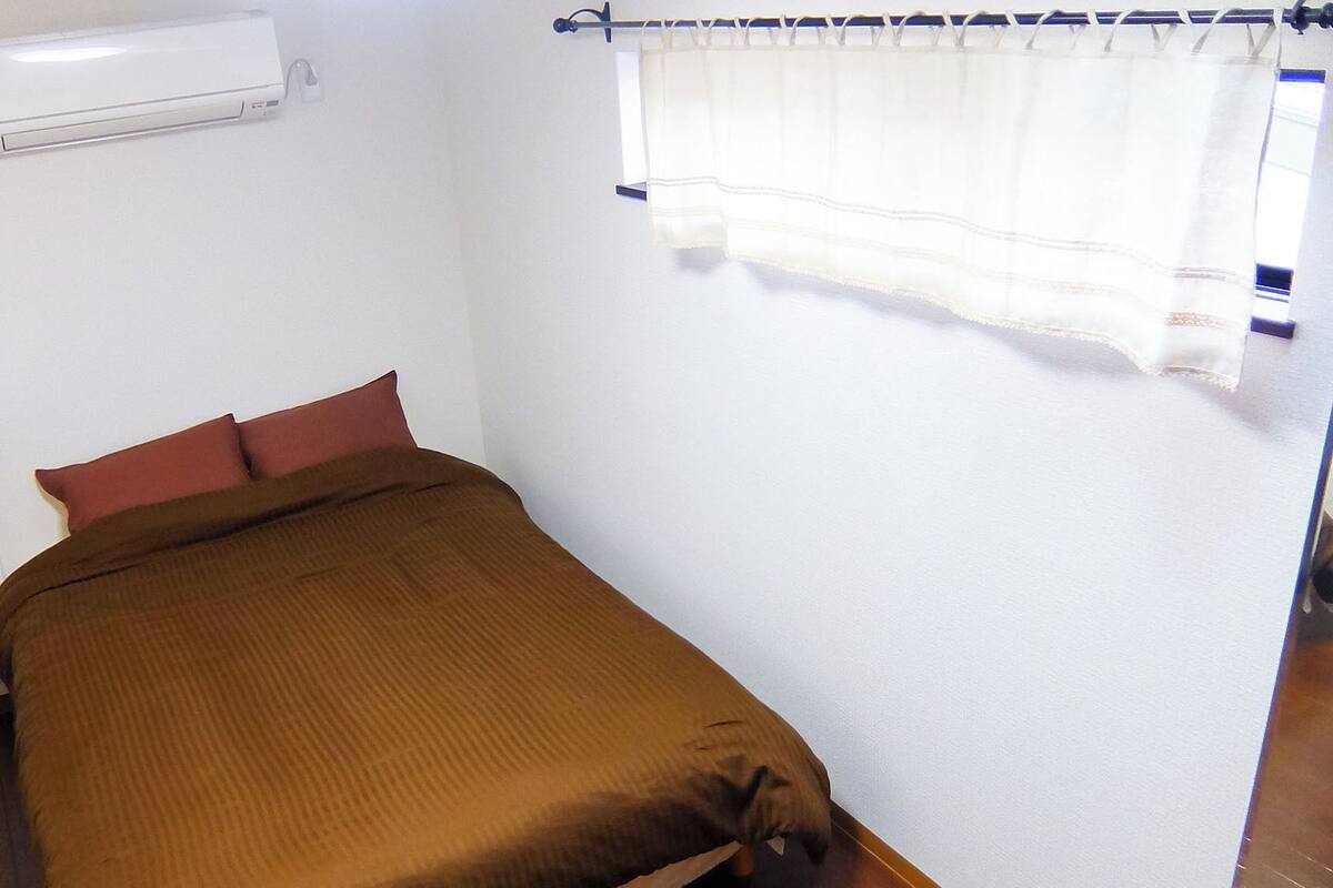 A single bed with a brown cover, a white air conditioner mounted on the wall, and a window with a white curtain.