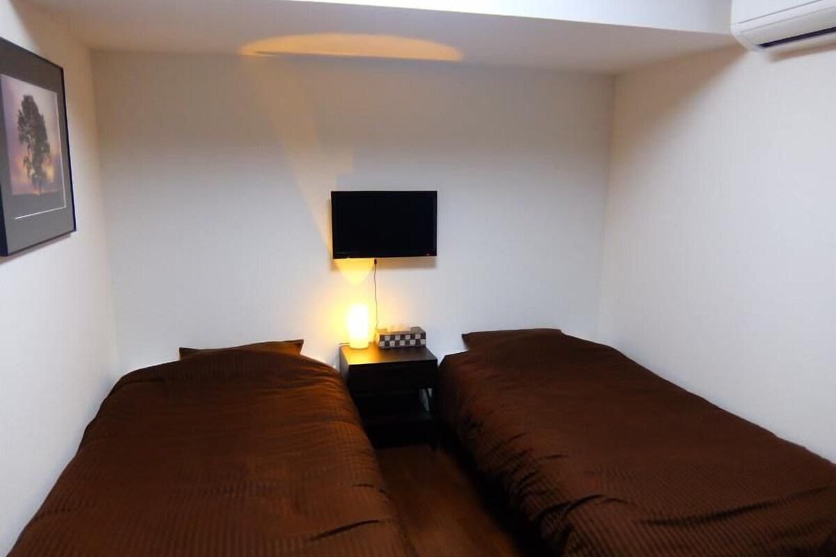 A single bed with a headboard, a bedside table with a lamp, and a flat-screen TV mounted on the wall.