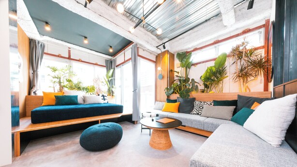 A modern living room with a sofa, ottoman, and coffee table, large windows with plants, and a visible ceiling with exposed beams. - Illi Nakano (Tokyo)