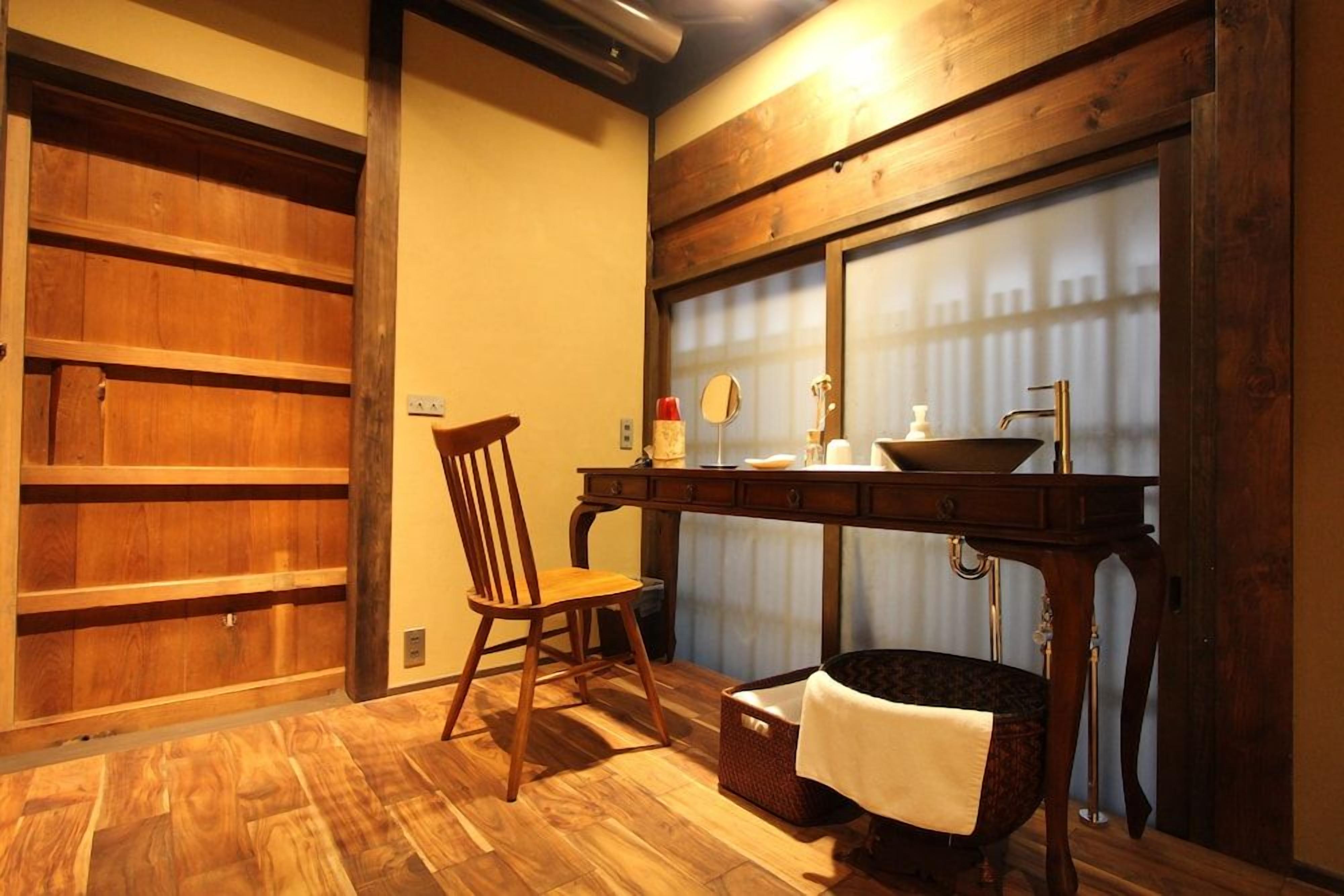 Una habitación con estructura de madera, una silla, una mesa con lavamanos y una ventana con persianas.