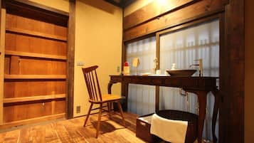 Una habitación con estructura de madera, una silla, una mesa con lavamanos y una ventana con persianas.