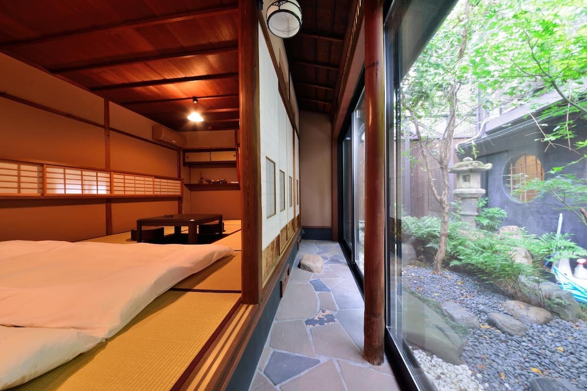 A traditional Japanese-style room with tatami flooring, a bed, a small table, and a view of a garden through a large window.
