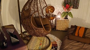Um quarto aconchegante com uma cadeira suspensa, um banco acolchoado, uma mesinha com luminária e uma planta em vaso.