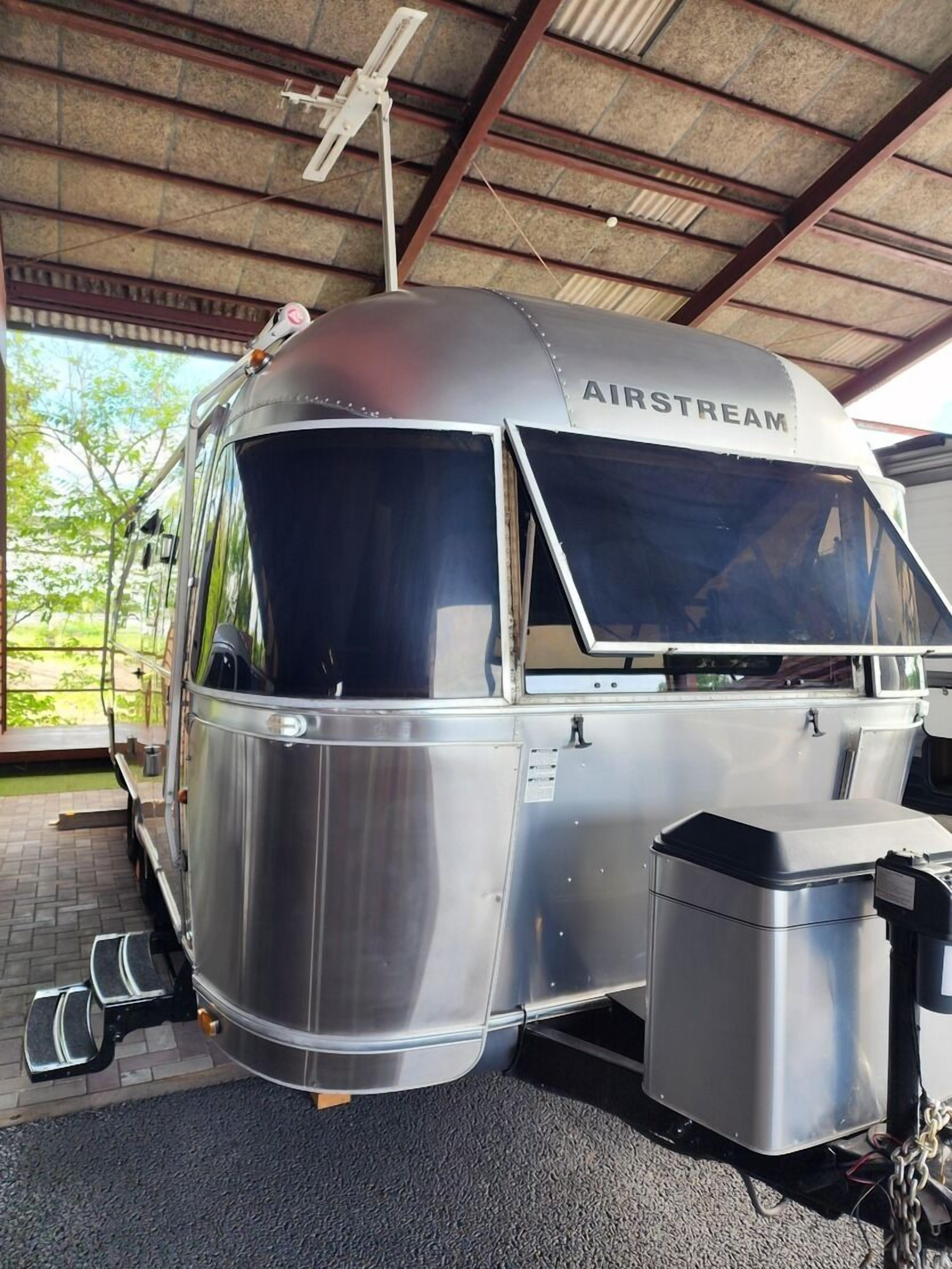 A stationary vehicle with a metallic exterior, labeled 'AIRSTREAM,' parked under a covered area with a roof and metal beams.