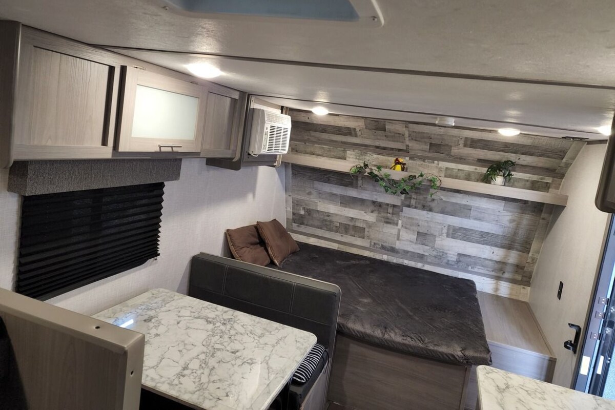 A compact RV interior with a kitchen area, a seating area with cushions, and a built-in shelf with plants.