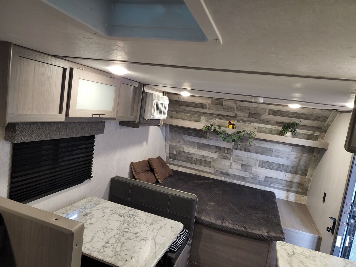A compact RV interior with a kitchen area, a seating area with cushions, and a built-in shelf with plants.