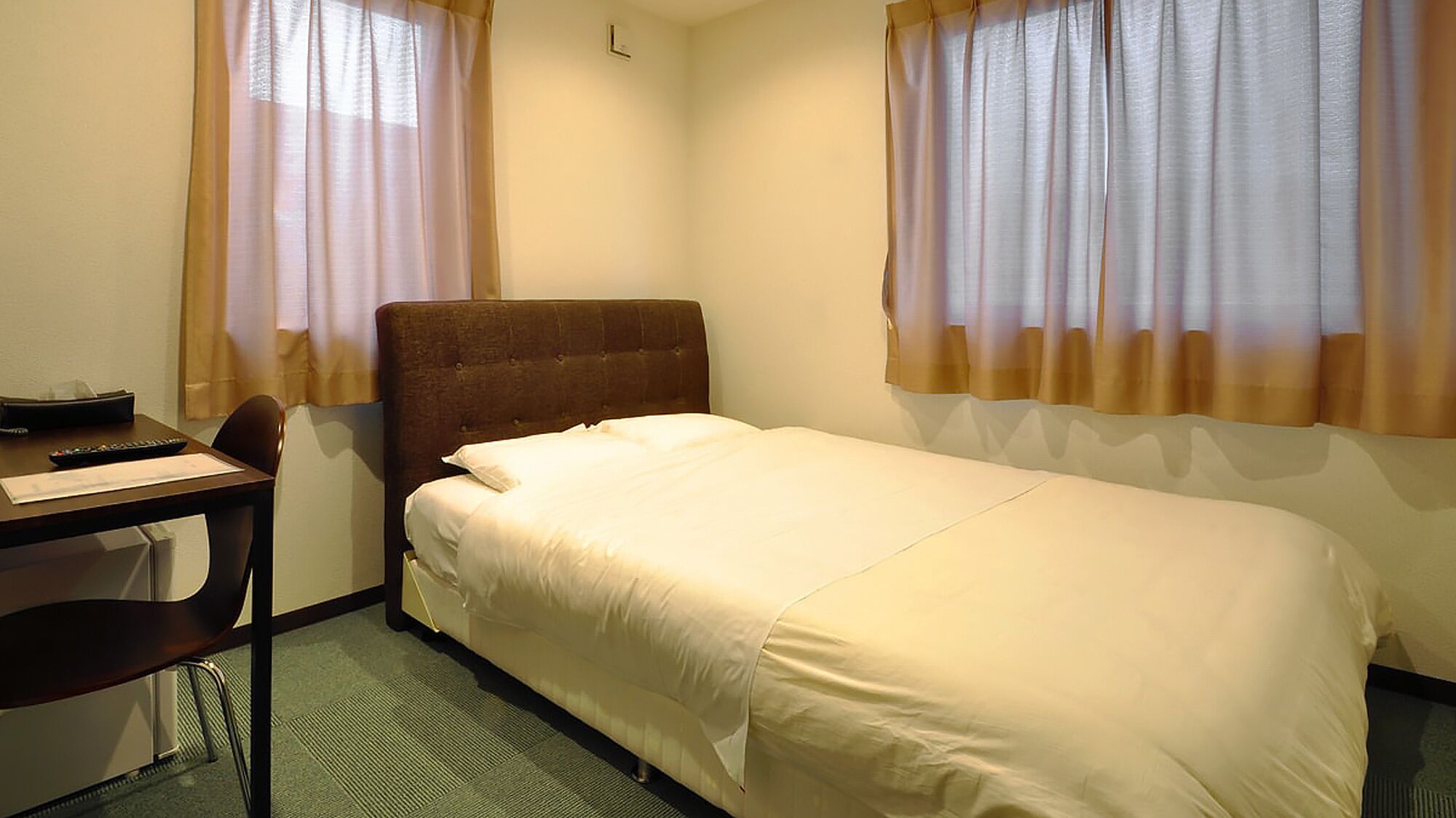 Ein Hotelzimmer mit einem Einzelbett, einem Schreibtisch mit Stuhl und zwei Fenstern mit durchsichtigen Vorhängen.
