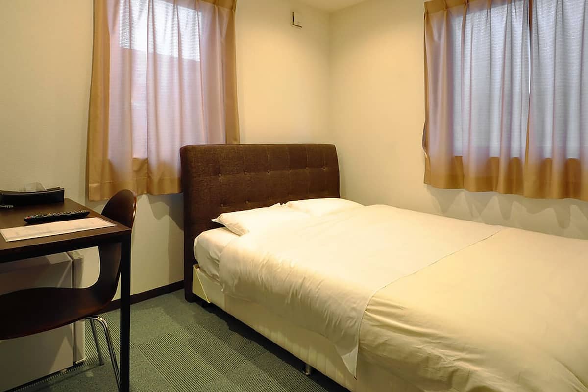 A hotel room with a single bed, a desk with a chair, and two windows with sheer curtains.