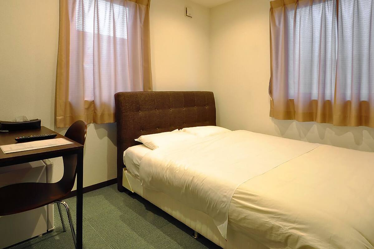 A single bed with a headboard, a desk with a chair, a remote control, and a pair of slippers on the floor.