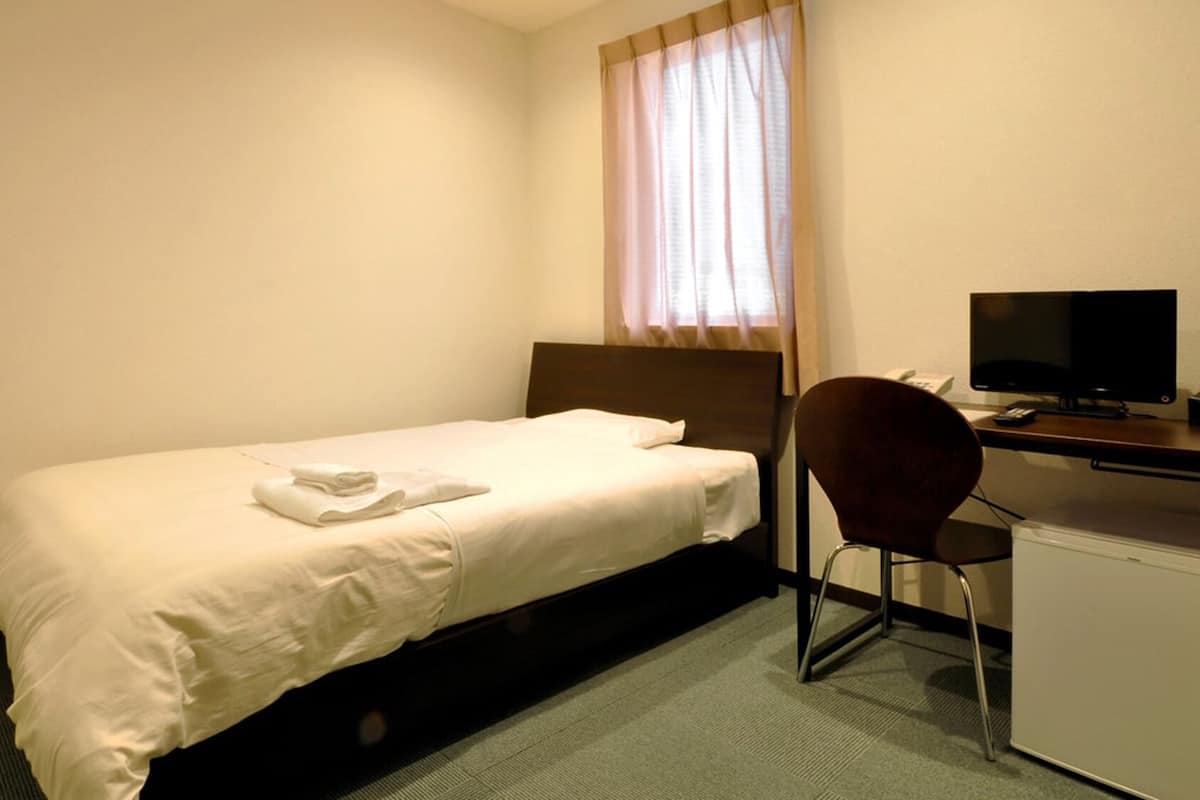 A single bed with a white comforter, a wooden headboard, a desk with a computer, a chair, and a small white cabinet.