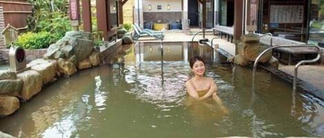 A person in a hot spring bath area with stone steps and a wooden structure overhead.