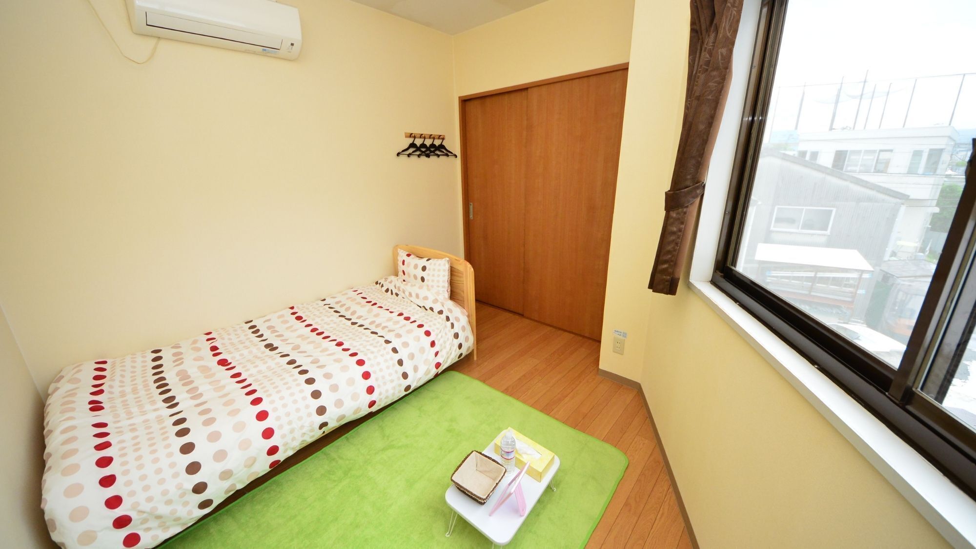 Ein kleiner, ordentlicher Raum mit einem Einzelbett, einer grünen Matte, einem Fenster mit Aussicht und einer Holztür.