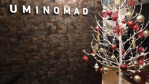 Un árbol de Navidad con adornos y luces, una pared de madera con el texto 'UMINOMAD' y un suelo.