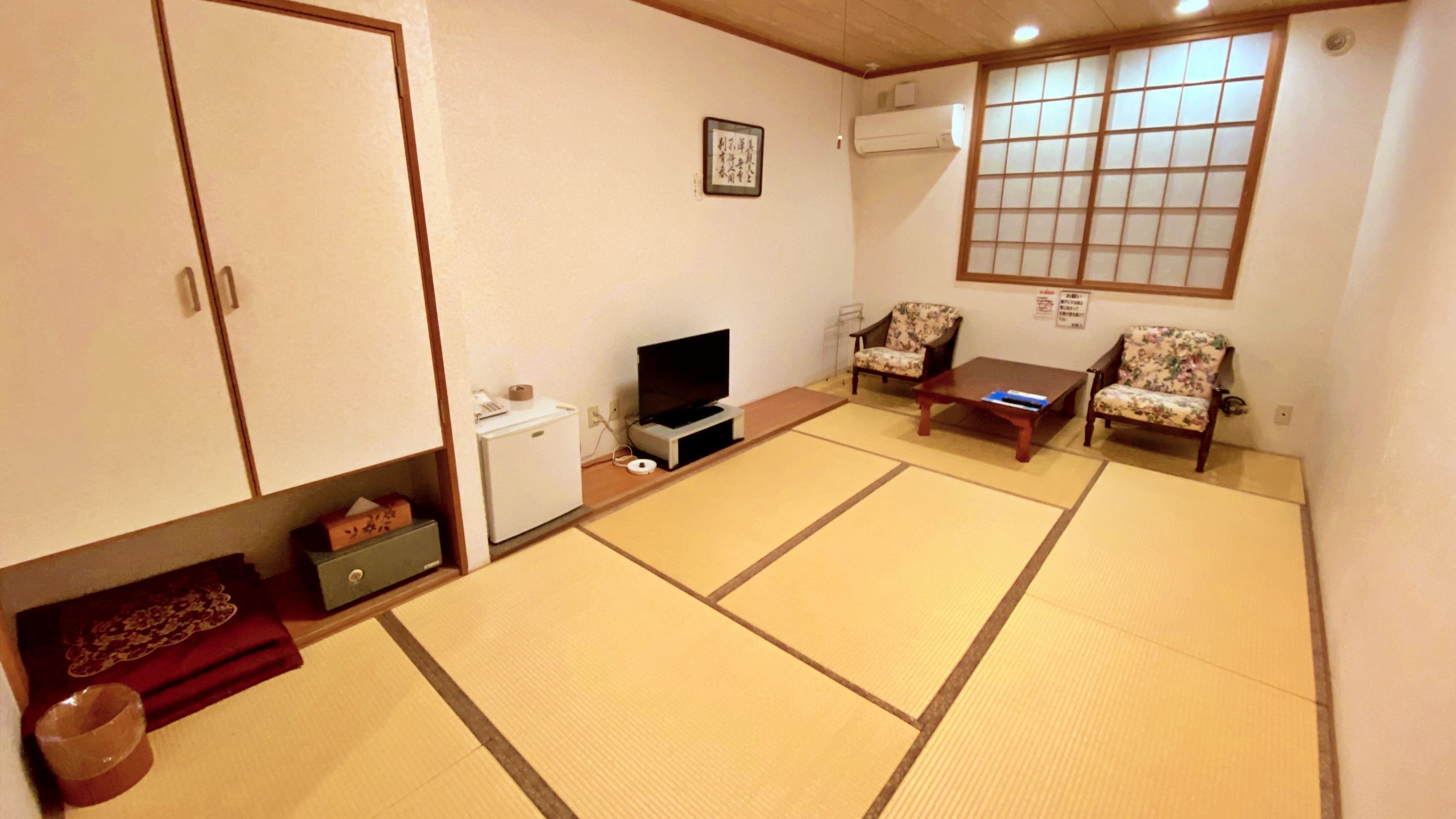 A traditional Japanese-style room with tatami flooring, a television, a small refrigerator, and two floral-patterned chairs.