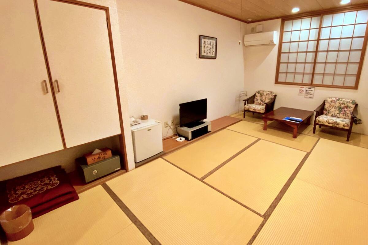 A traditional Japanese-style room with tatami flooring, a television, a small refrigerator, and two floral-patterned chairs.