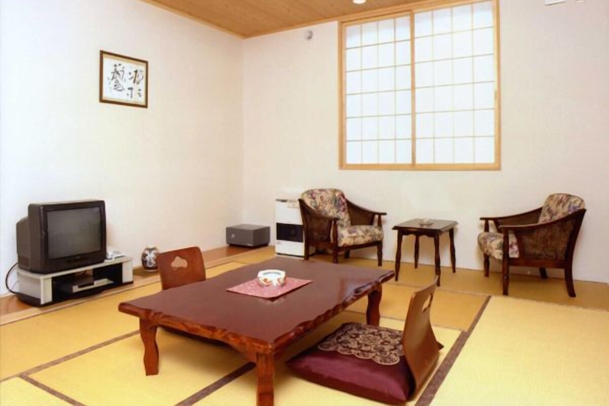 Une pièce de style japonais traditionnel, avec un sol recouvert de tatamis, une table basse, deux chaises, un téléviseur et une grande fenêtre.