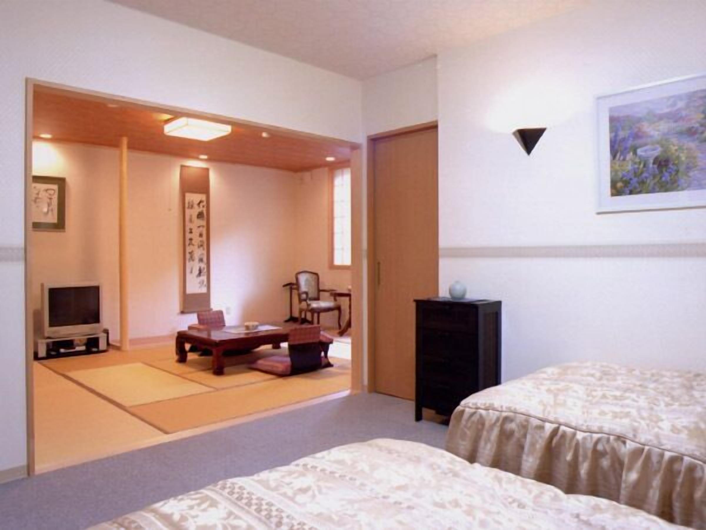 A room with tatami mats, a low table, chairs, a TV, a bed, and a painting on the wall.