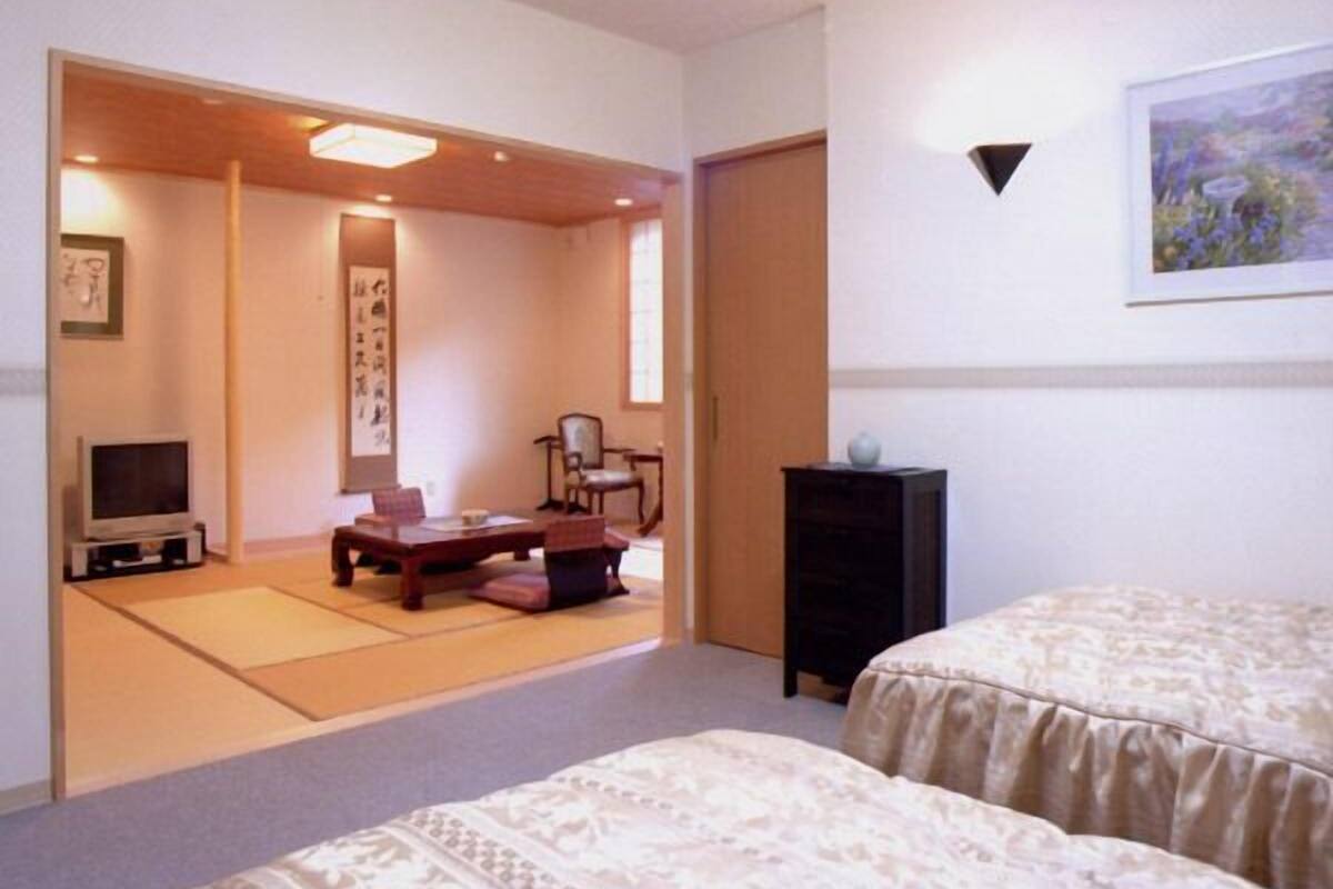 A room with tatami mats, a low table, chairs, a TV, a bed, and a painting on the wall.