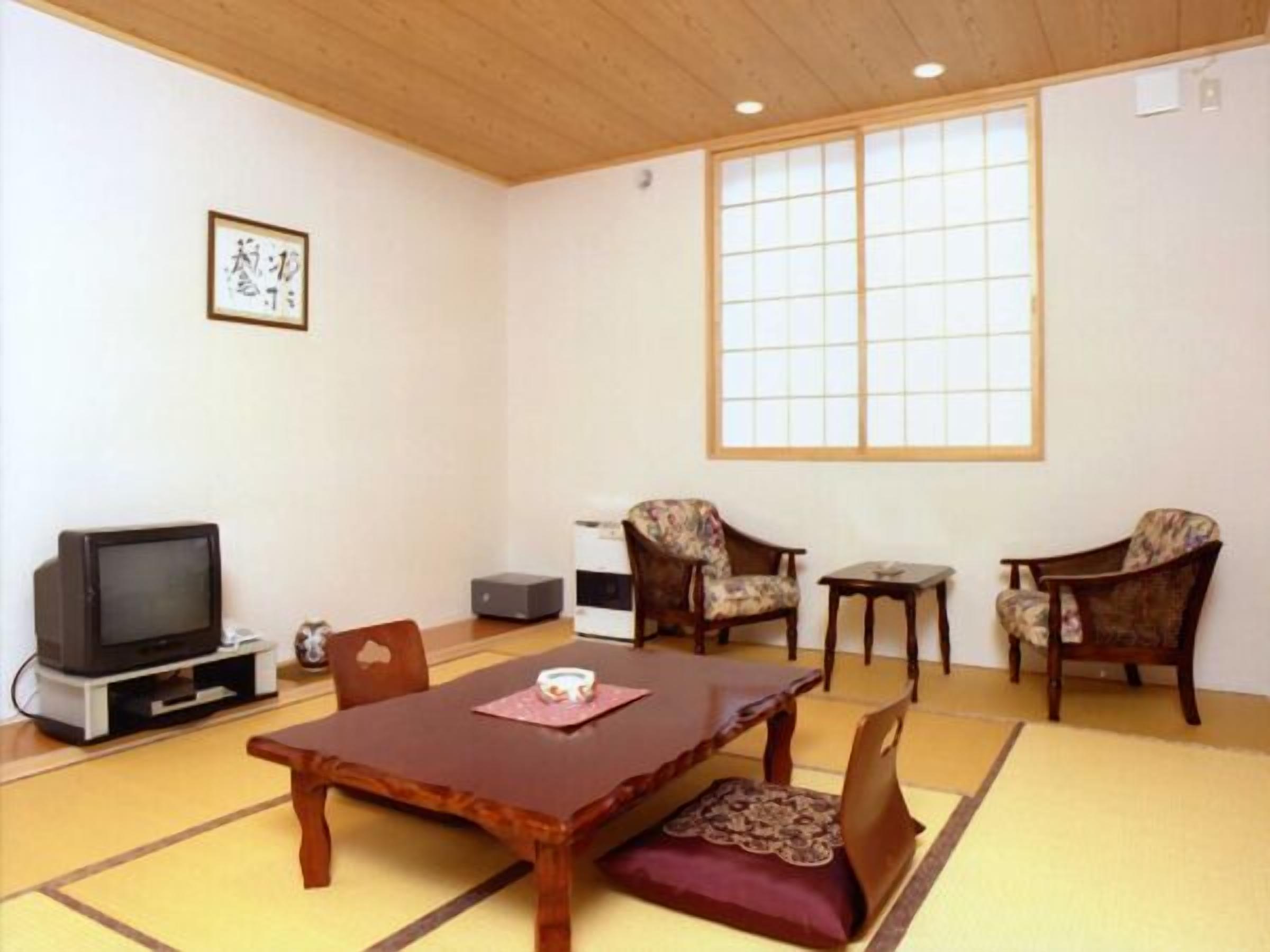 A traditional Japanese room with tatami flooring, a low table, two chairs, a television, and a large window.