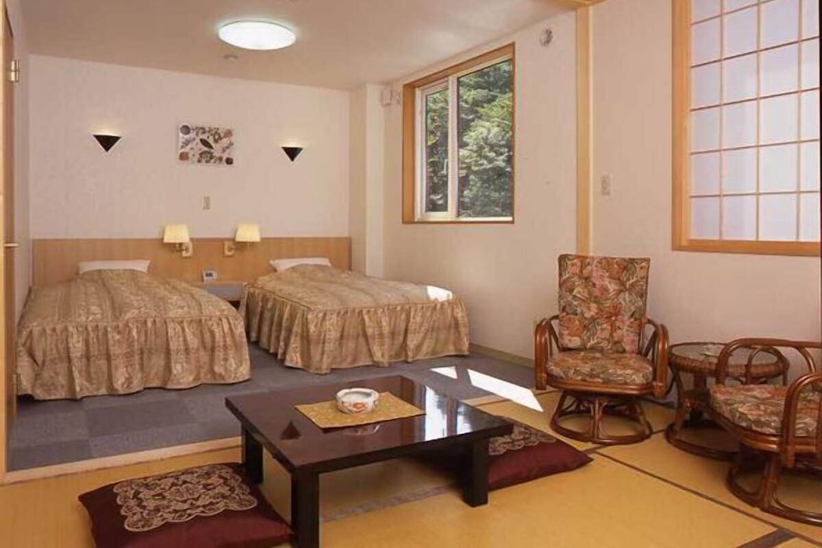 A hotel room with two beds, a small table, a chair, and a window.