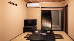 Quarto com piso de tatame, televisão sobre um suporte, porta deslizante e uma pequena mesa com xícaras e uma revista.