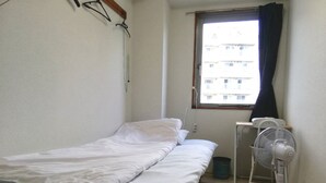 Una camera piccola con un letto singolo, una finestra, un ventilatore e un cestino.