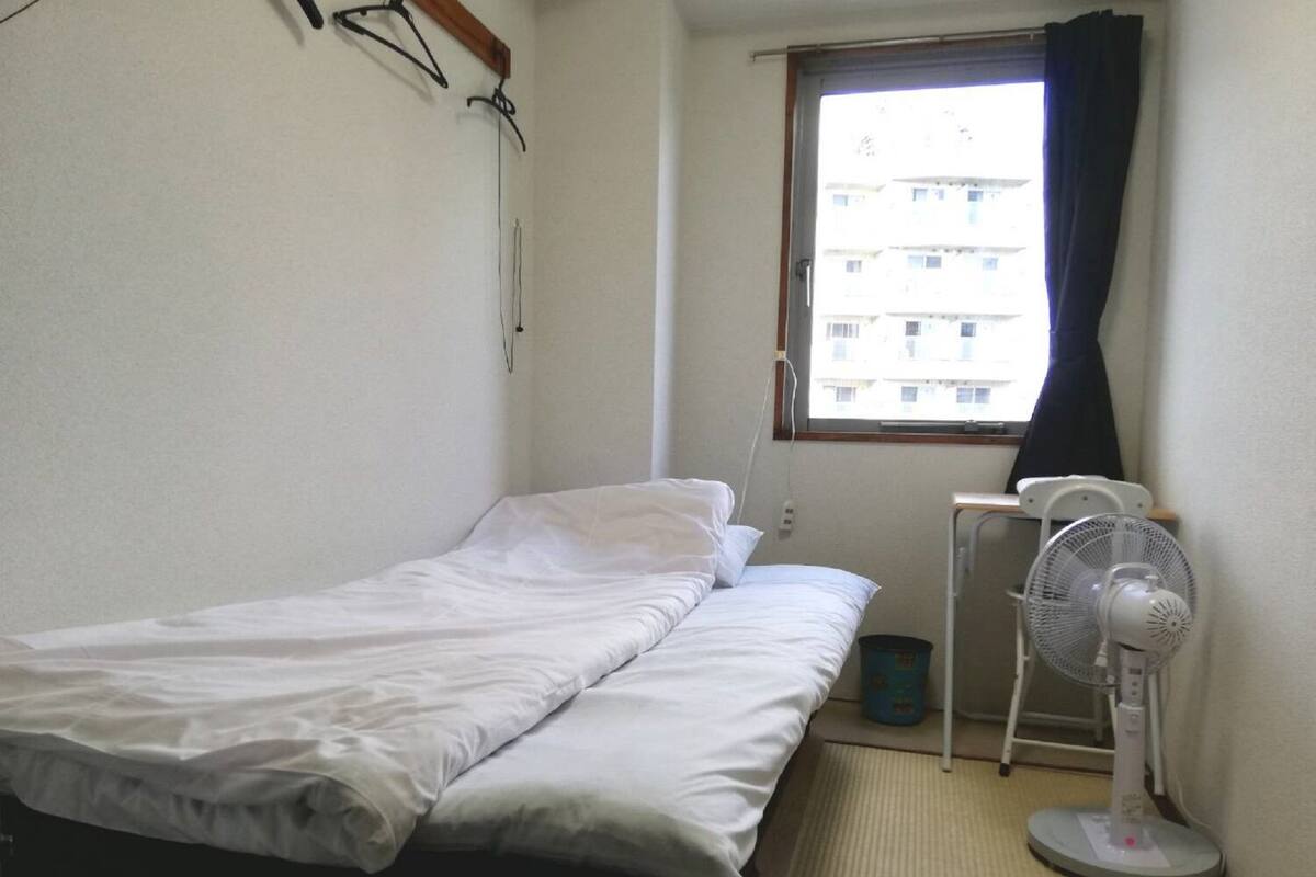 A small, single-bed room with a window, a standing fan, and a waste bin.